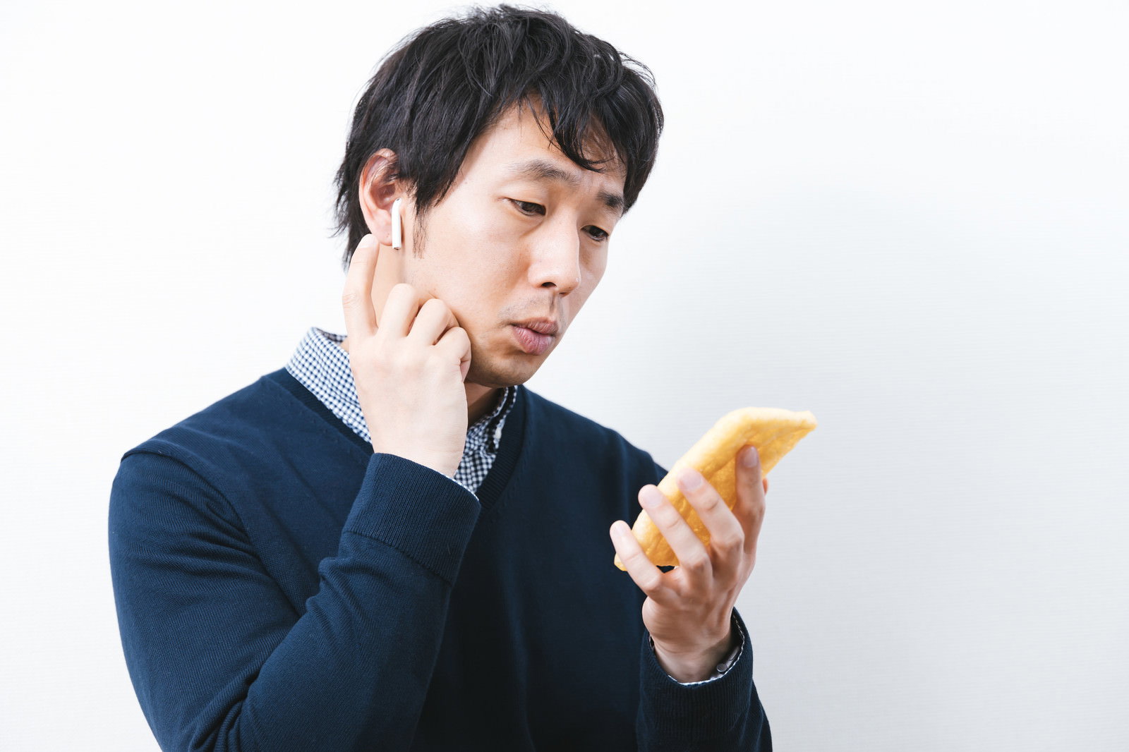 ワイヤレスイヤホンを装着しながら油揚げを持って困惑している男性［モデル：大川竜弥］