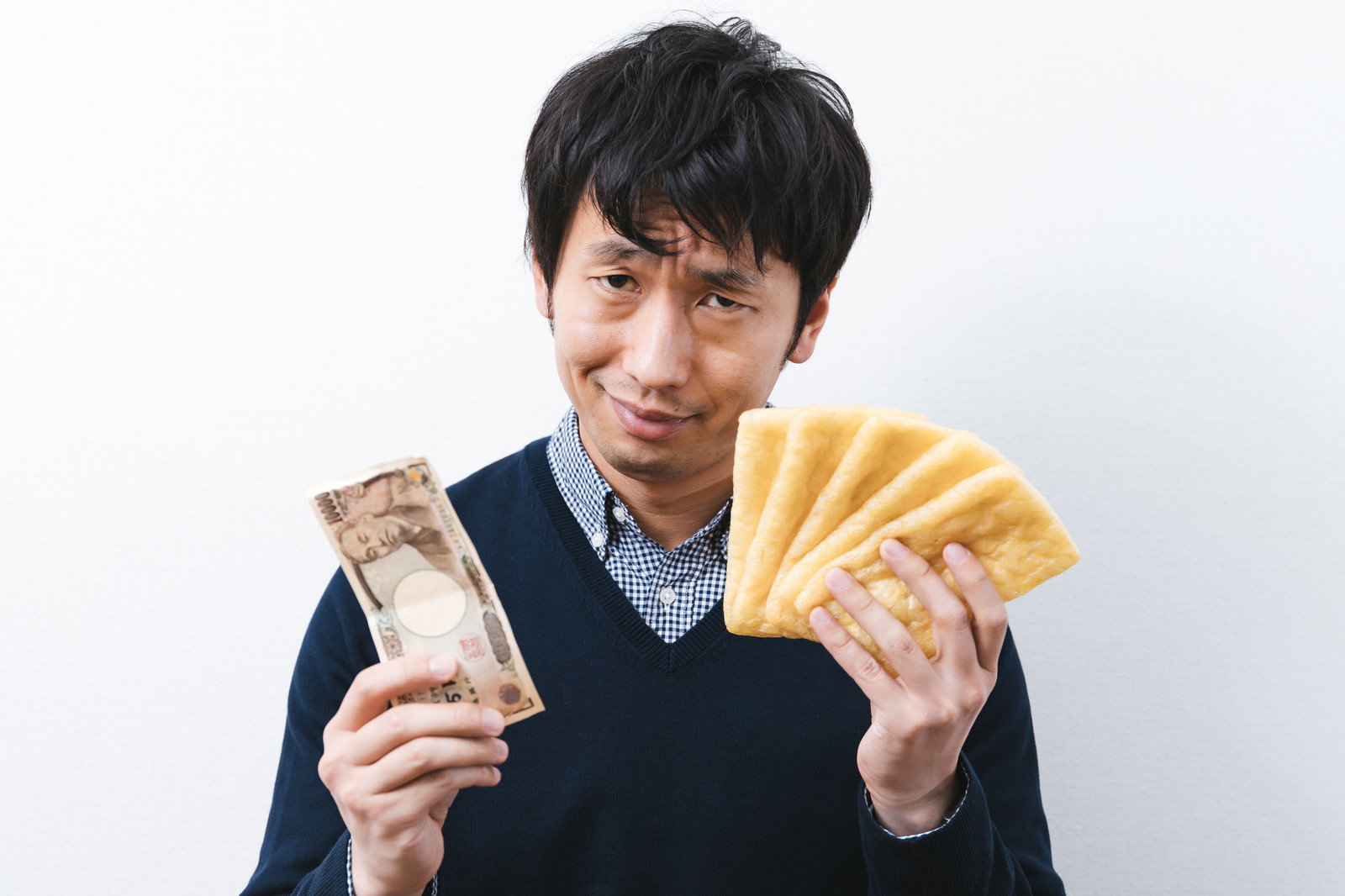 油揚げと一万円札を両手に持って笑顔を見せる紺色のベストを着た男性［モデル：大川竜弥］