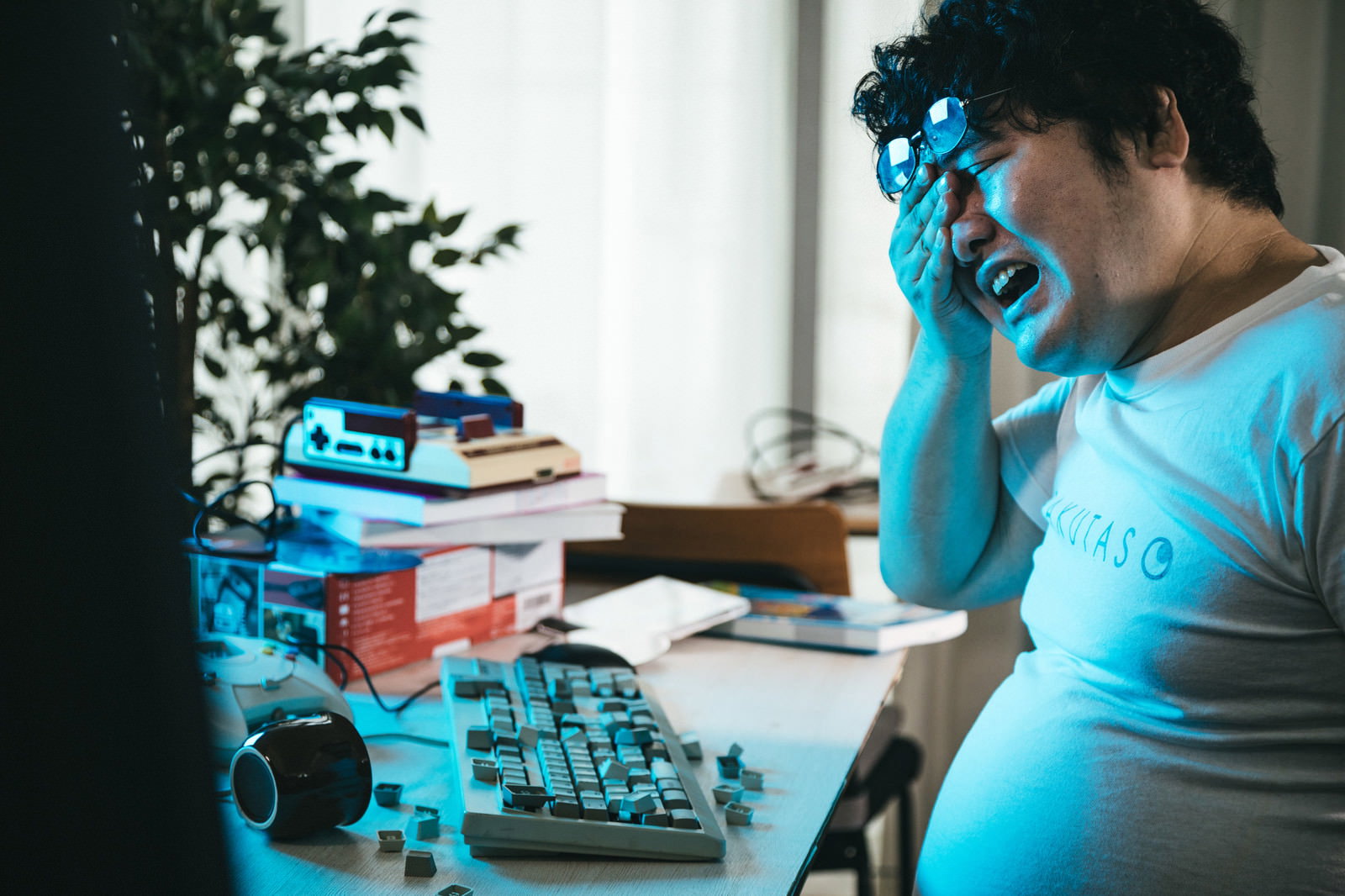 青い照明下で両手で顔を覆う男性がデスクに座っている。キーボードと書類が机上にある［モデル：段田隼人］