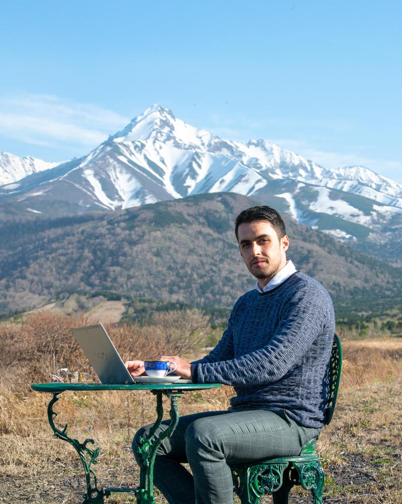 雪山を背景にノートパソコンで作業するディレクター男性［モデル：アドリアーノ］