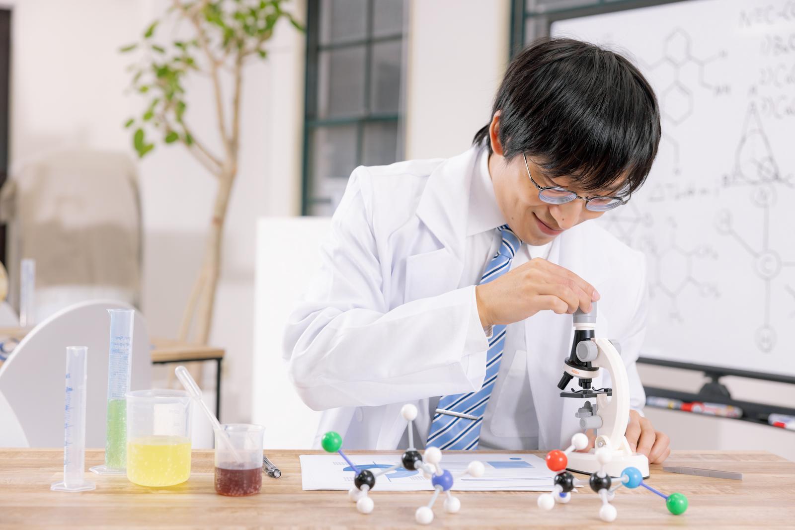 白衣を着た男性が顕微鏡で観察実験をしており、テーブルには試験管と分子模型が置かれている［モデル：あづま］