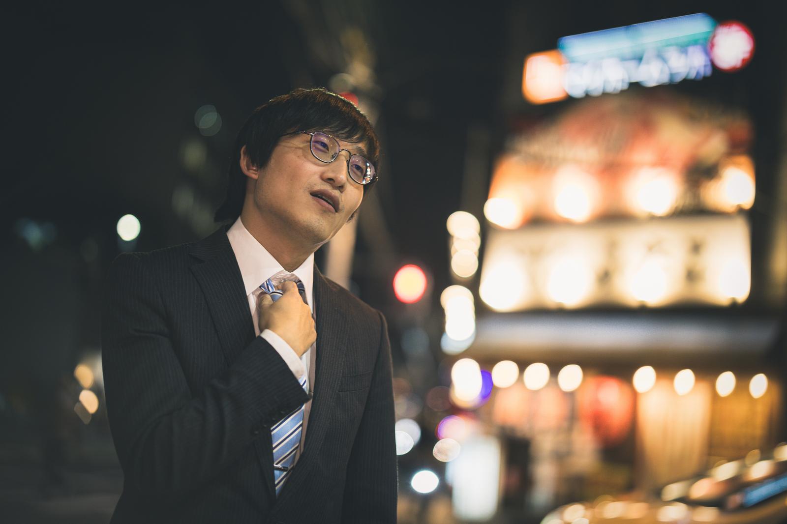 ネクタイを直すスーツ姿の男性が夜間の街で立っている［モデル：あづま］
