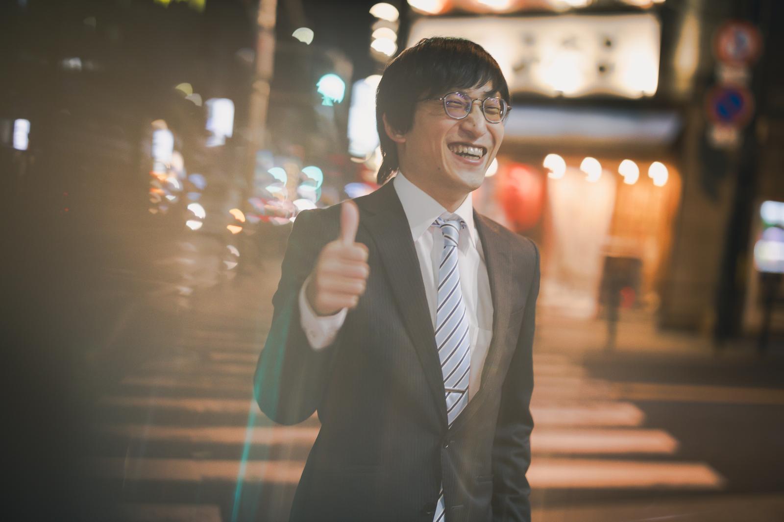 夜間の繁華街で親指を立てて笑うスーツ姿の酔っぱらった会社員男性［モデル：あづま］