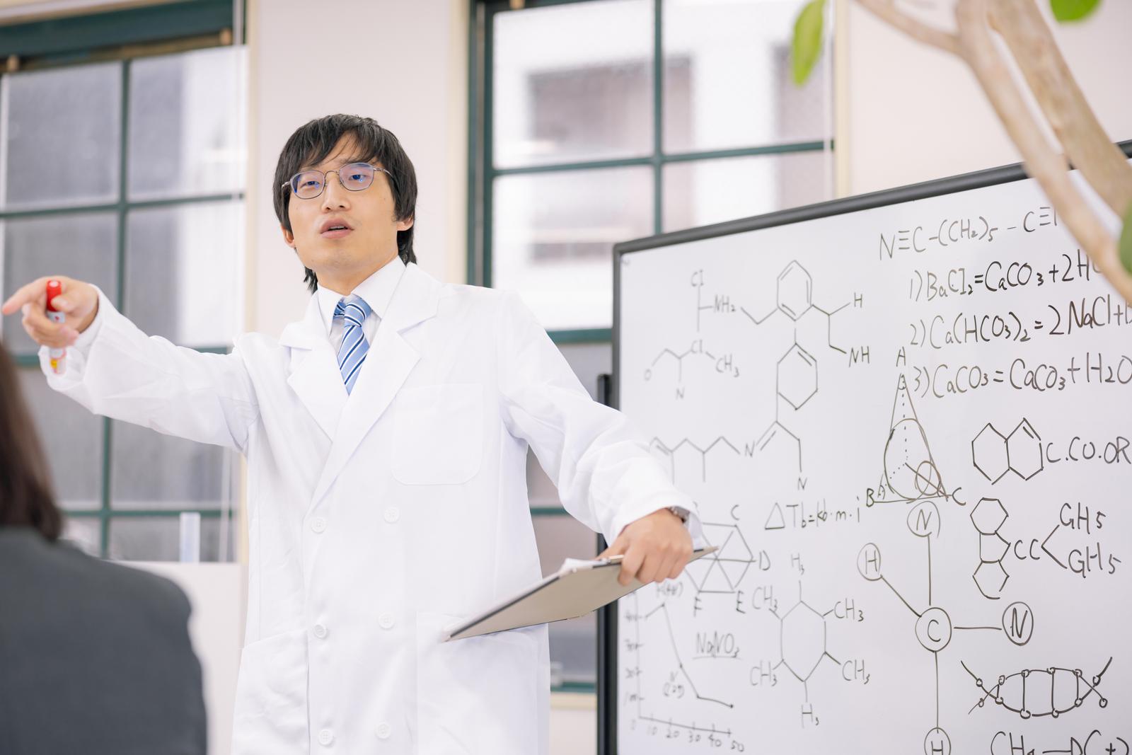 ホワイトボードに化学式を書きながら講義する白衣姿の男性科学者［モデル：あづま］