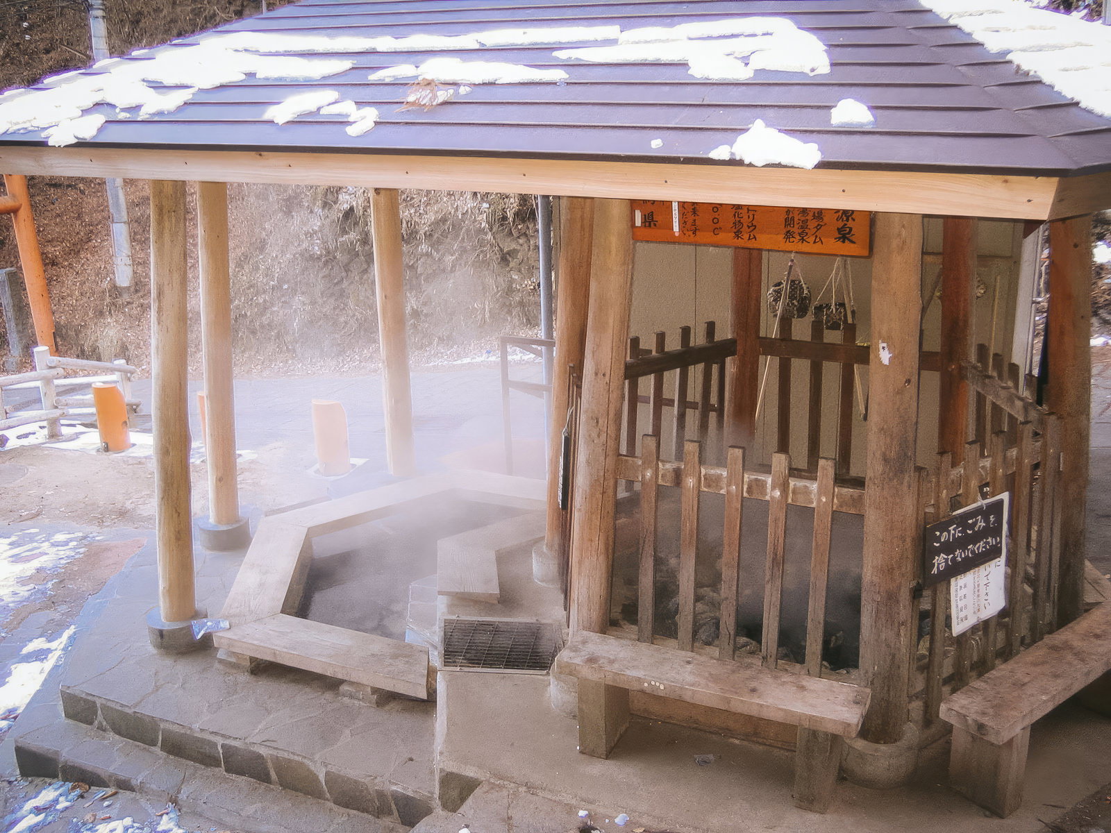 白い雪が積もった木造屋根付き足湯施設。湯船から湯気が立ち上り、奥にはガラス張り待合所が見える冬景色