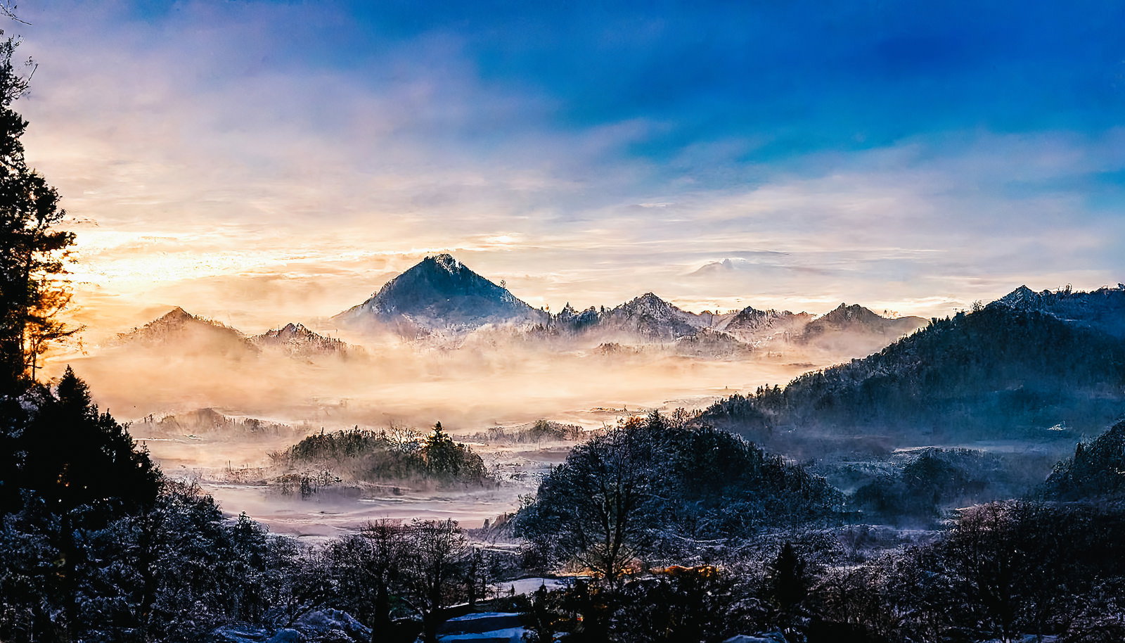 雪化粧を纏った山脈の間に朝霧が流れ、初日の出の朝日が照らす冬の風景