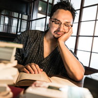 古民家の和室で積読に囲まれて読書に集中する男性の写真