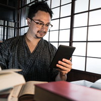 古民家の畳の上で積まれた本よりタブレットで電子書籍を読む男性の写真