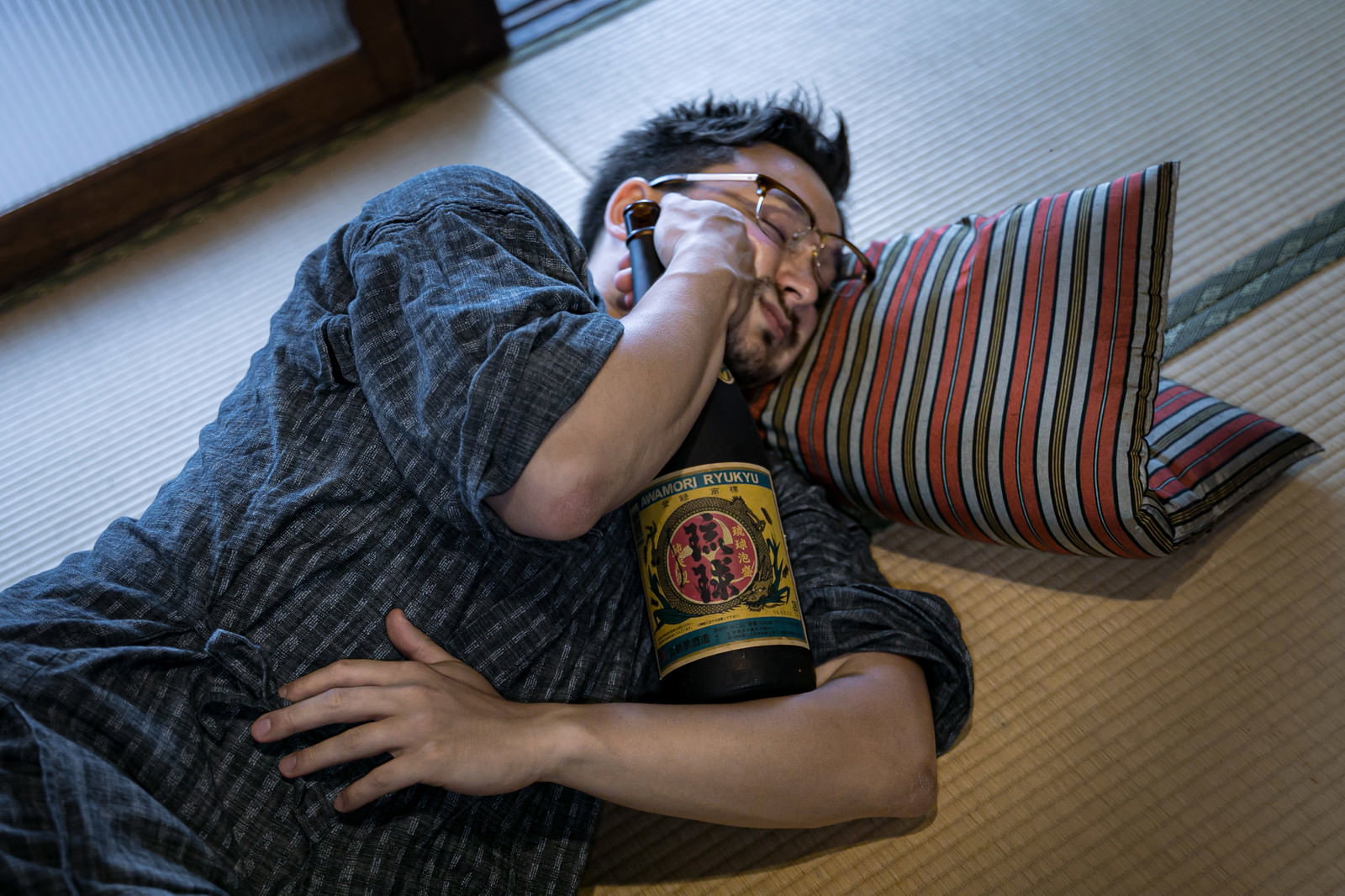 畳の上で一升瓶を抱えて泥酔し横たわるメガネの男性［モデル：Max_Ezaki］