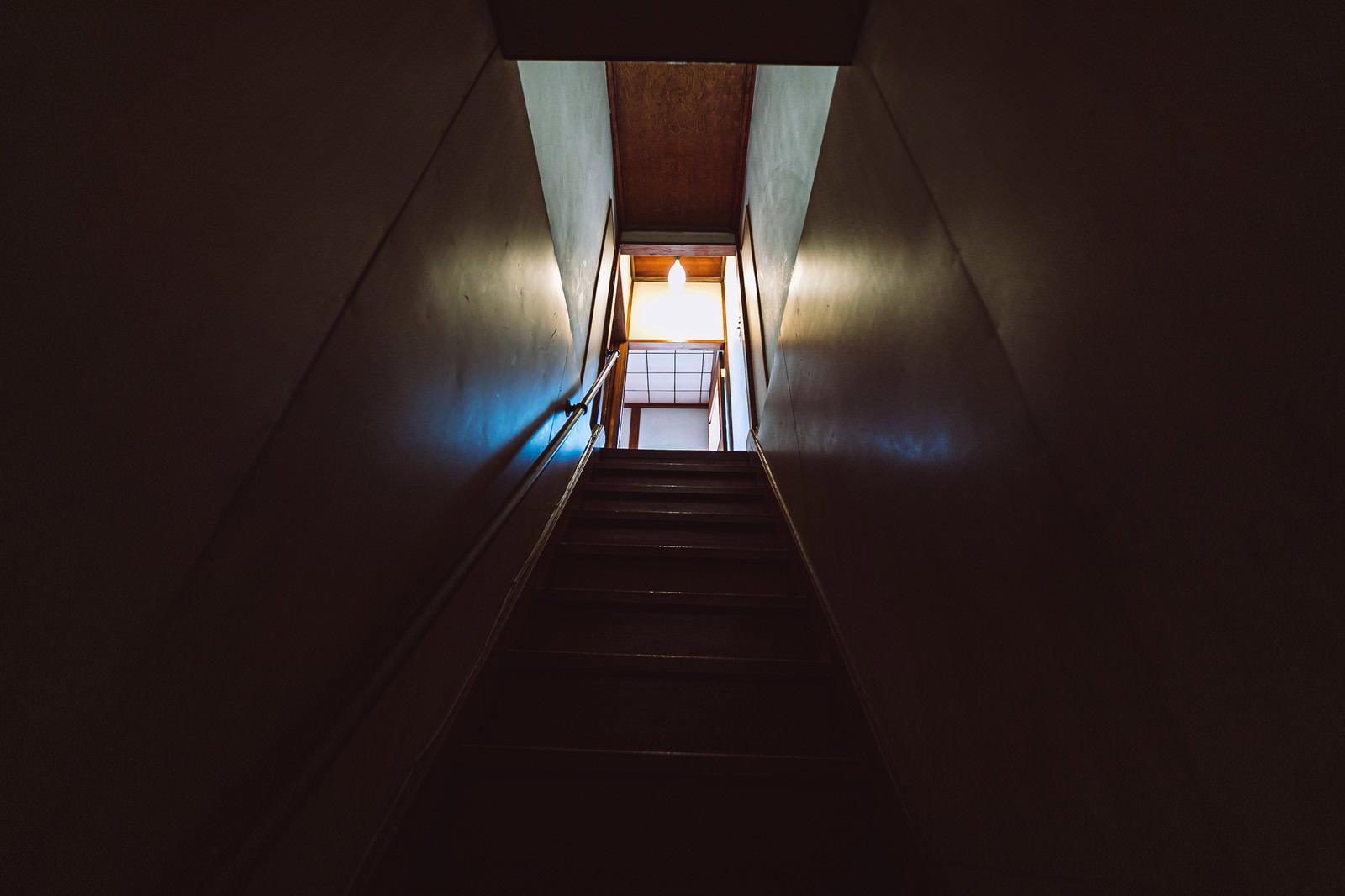 薄暗い古民家の室内で、上階から光が差し込む木造の階段