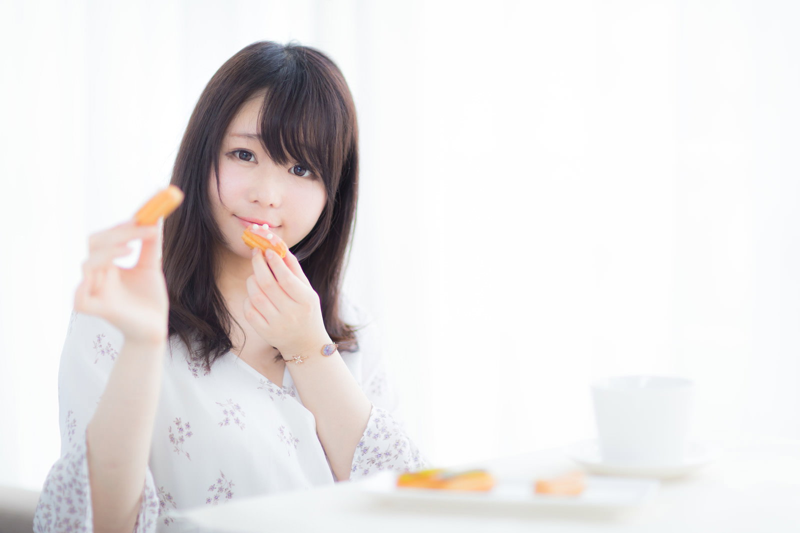 エクレアを手に持ち笑顔を見せる白い服の女性［モデル：茜さや］