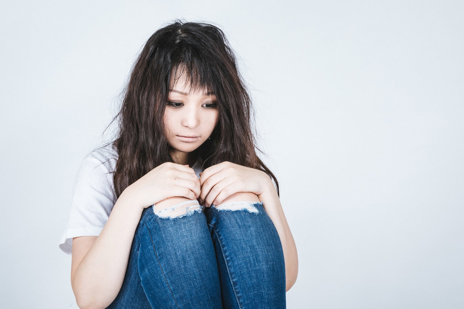 白い長袖とデニムを着た女性が体育座りでうなだれている様子［モデル：茜さや］
