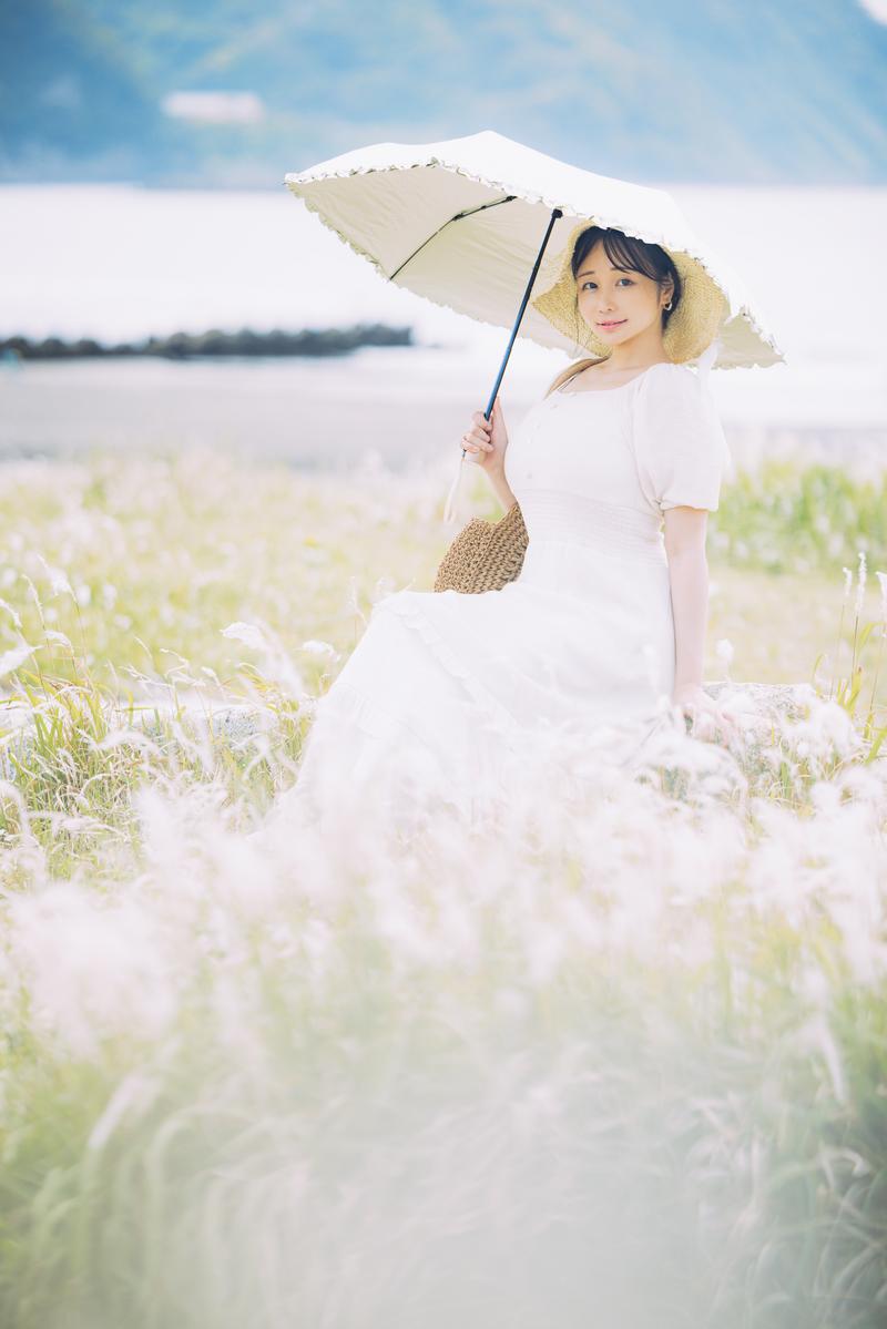 河川敷の草地に立つ女性が麦わら帽子と白いドレスで日傘をさしている［モデル：茜さや］