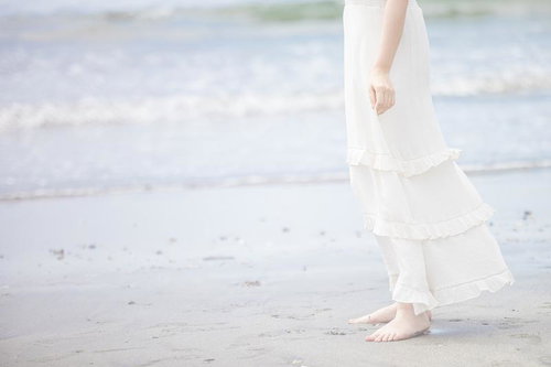 波打ち際で裸足の白いワンピース姿の女性が立つ浜辺の風景
