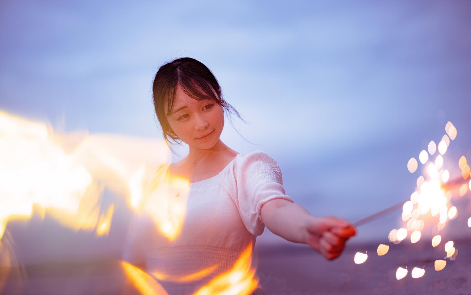 夜間にオレンジ色の火花が散る手持ち花火を持つ女性のポートレート写真［モデル：茜さや］