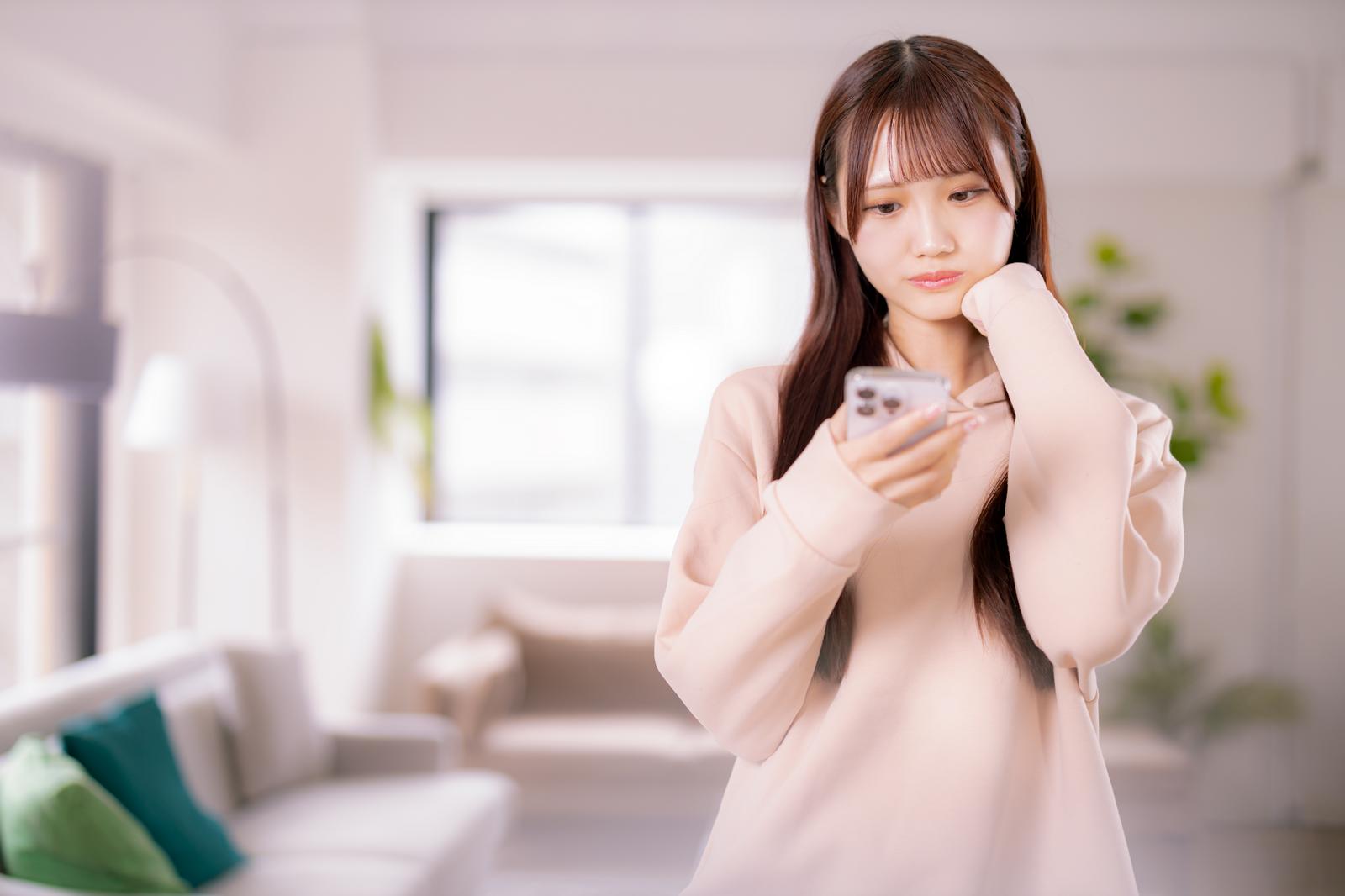 スマホを持ちながら考え事をする女性が、明るい室内で壁に寄りかかっている［モデル：あき］
