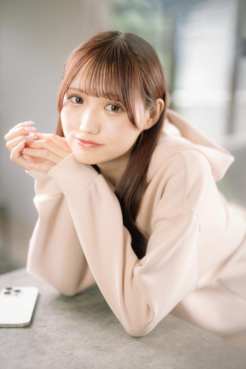 朝の光の中でベージュニットを着た女性がスマートフォンを持ち微笑んでいる［モデル：あき］