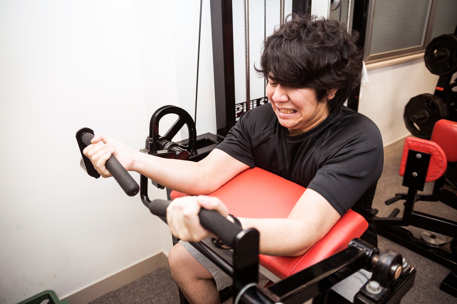赤いシートの器具でバイセップスカールをする男性が必死の表情で二頭筋トレーニングに取り組んでいる［モデル：あまのじゃく］
