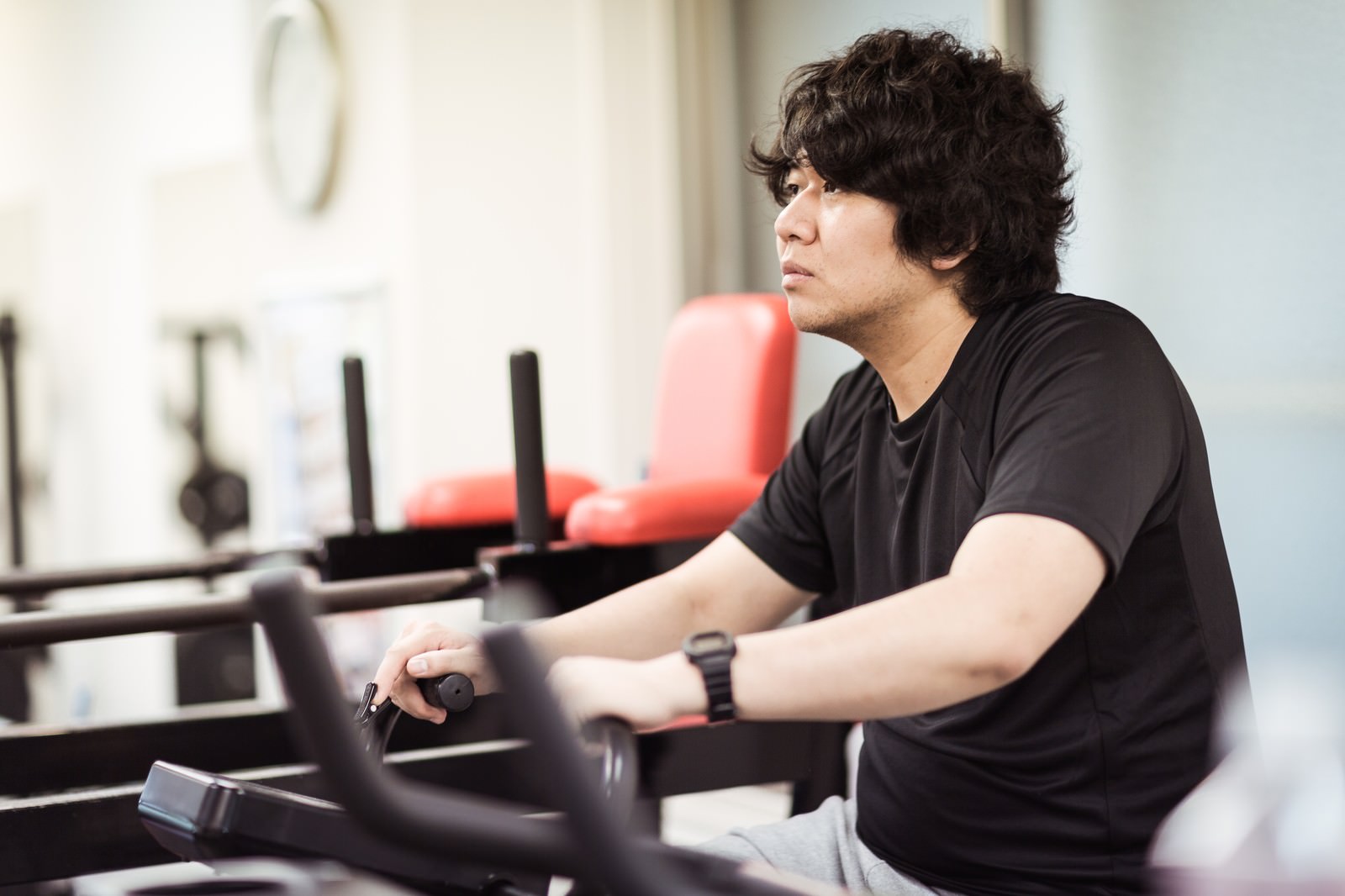 ジムでエアロバイクに乗りながらテレビを見る黒いTシャツの男性［モデル：あまのじゃく］