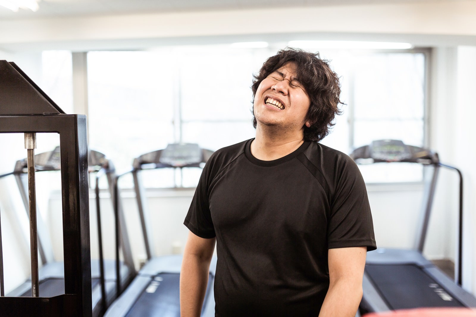 黒いTシャツを着た男性がランニングマシンで目を閉じ、辛い表情をしている様子。背景にジム器具［モデル：あまのじゃく］