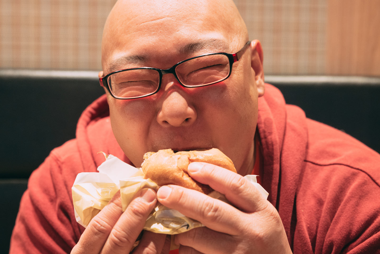 眼鏡をかけた男性が赤いシャツでハンバーガーを食べている［モデル：あまやん］