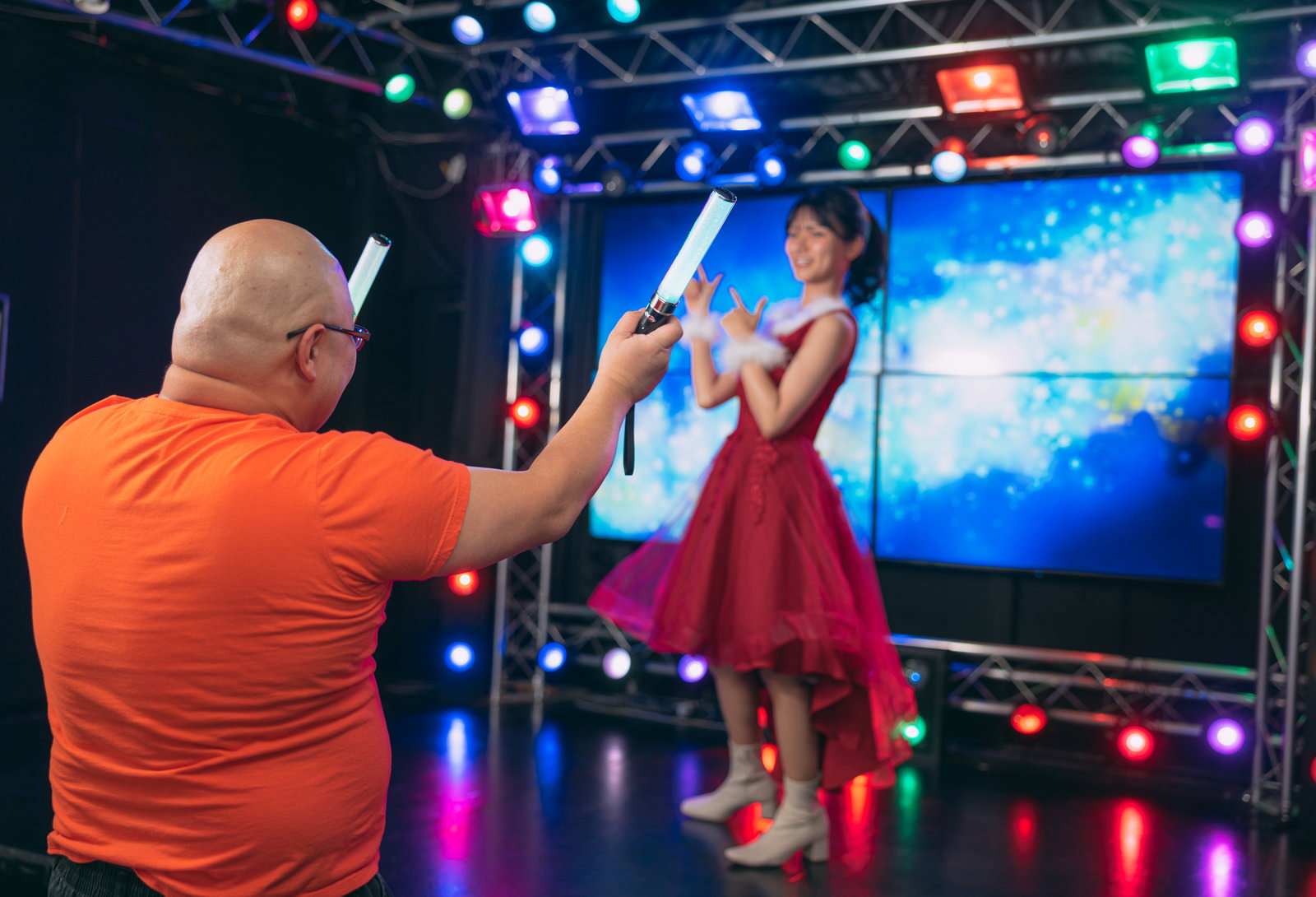 ステージ上のアイドルに向けてペンライトを掲げるオレンジ色のシャツを着た男性ファン［モデル：あまやん モデルリリース］
