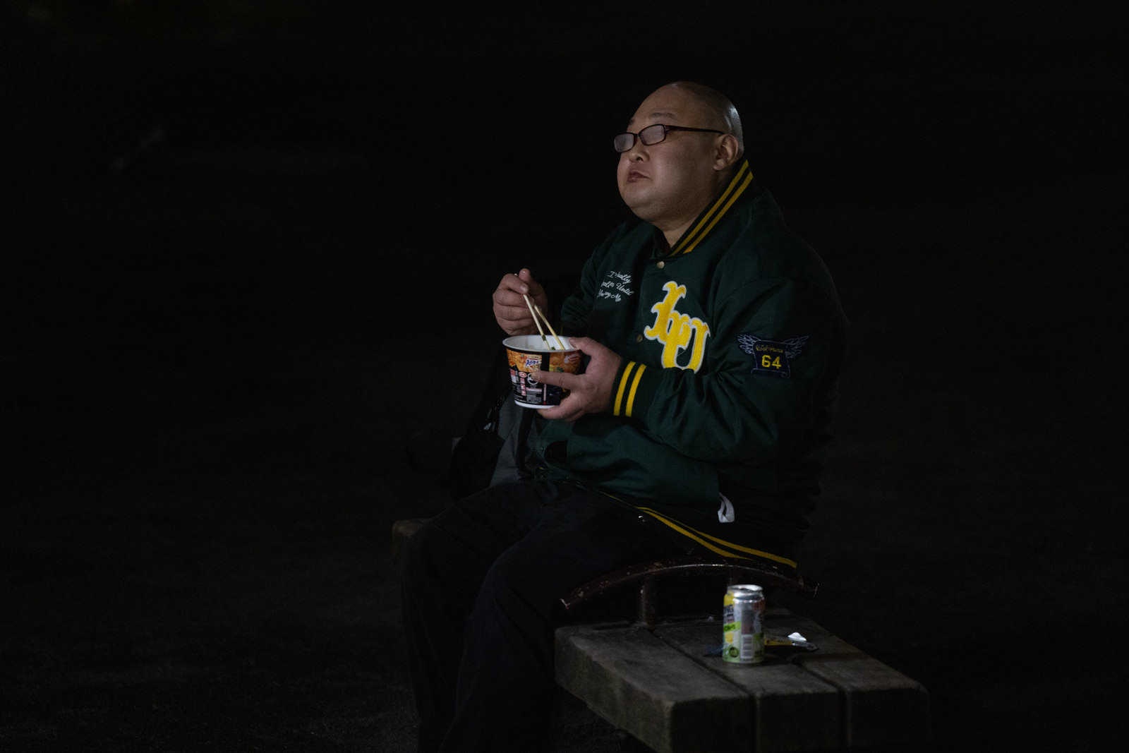 暗い背景の中、カップ麺を食べて眼鏡が曇る緑色のジャージを着た男性［モデル：あまやん］