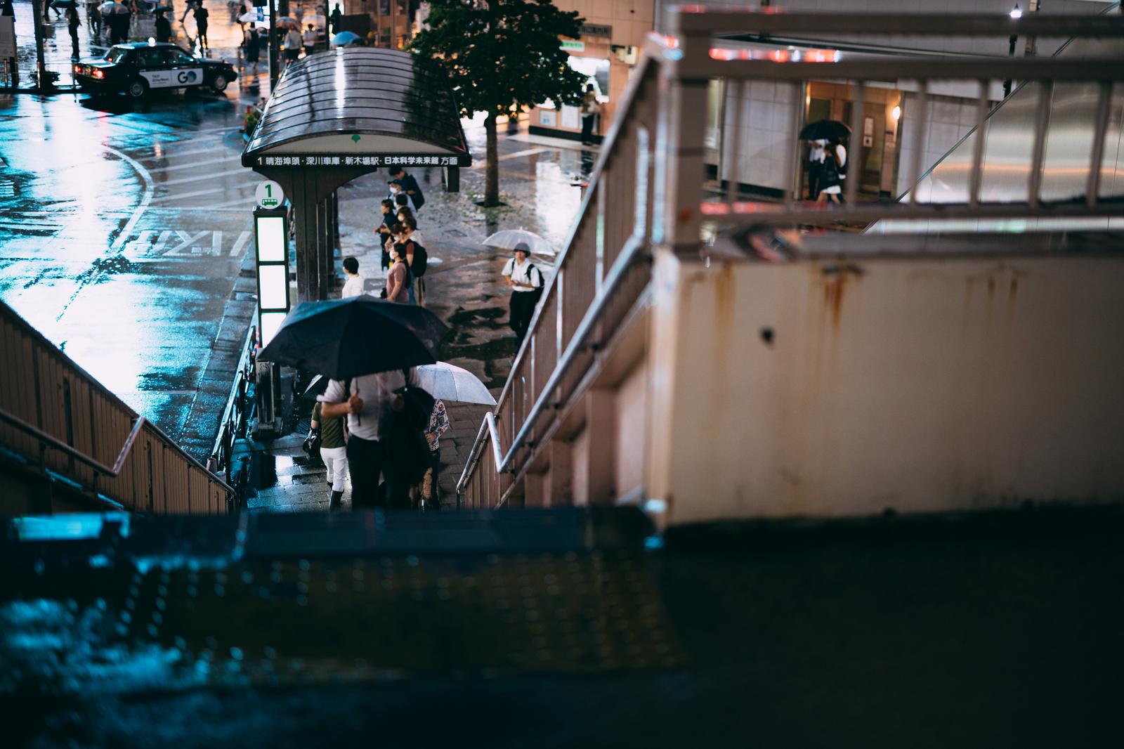 雨上がりの夜間、歩道橋の上からバス停で傘を差す人々を見下ろした写真