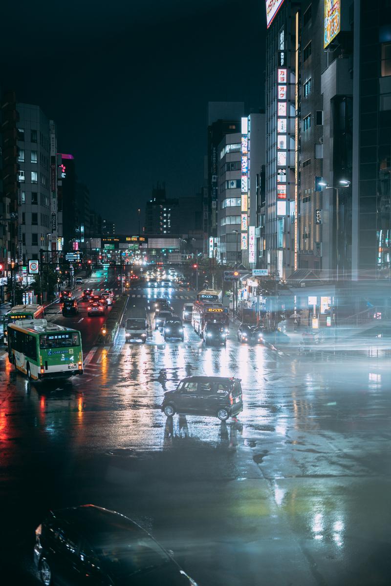 雨で濡れた夜の交差点、ネオンライトと車のライトが映り込む路面