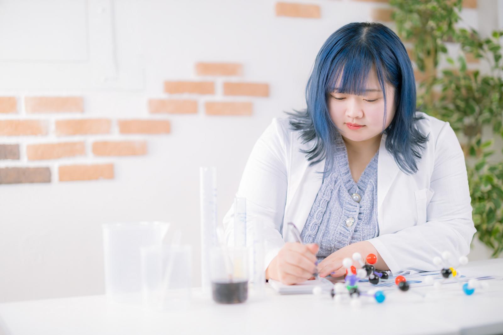 白衣の青髪女性が分子模型の前で記録をとる［モデル：あねちゃん］