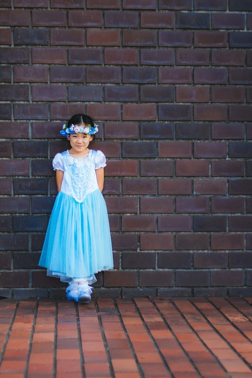 白い花飾りをつけた青いドレスの女の子がレンガ壁の前で笑顔