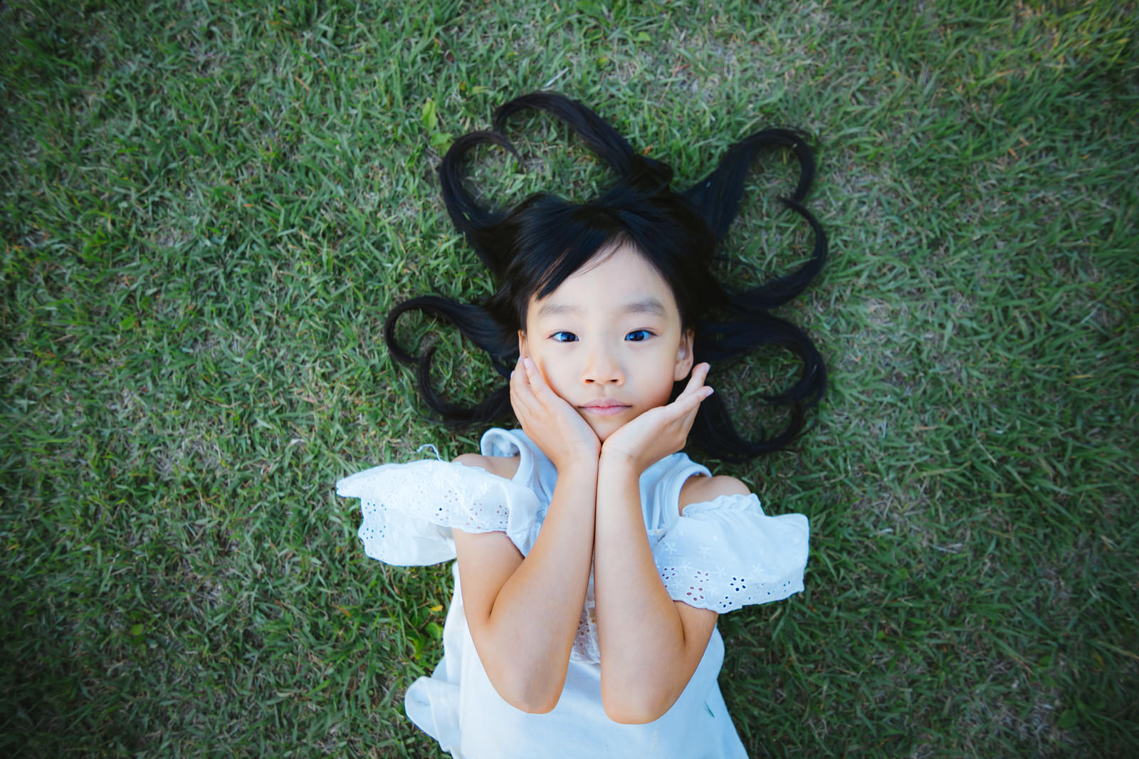 緑の芝生の上で白いワンピースを着た女の子が頬杖をついて寝転んでいる様子［モデル：あんじゅ］