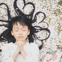 桜の花びらに横たわり両手を合わせて祈る女の子の写真
