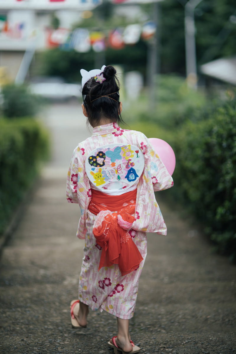 カラフルな浴衣を着た少女が赤い団扇を手に持って歩く後ろ姿［モデル：あんじゅ］