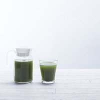 濃い緑色の青汁が入ったポットとグラス、白い背景の写真
