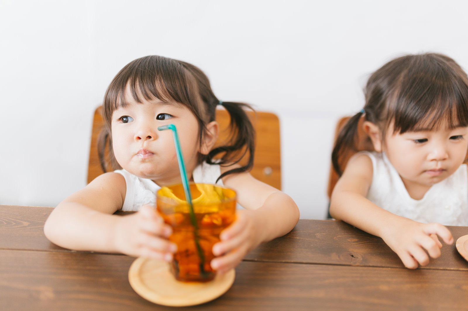 オレンジジュースのストローを咥える双子の女の子が木製テーブルに座っている［モデル：あおみどり］