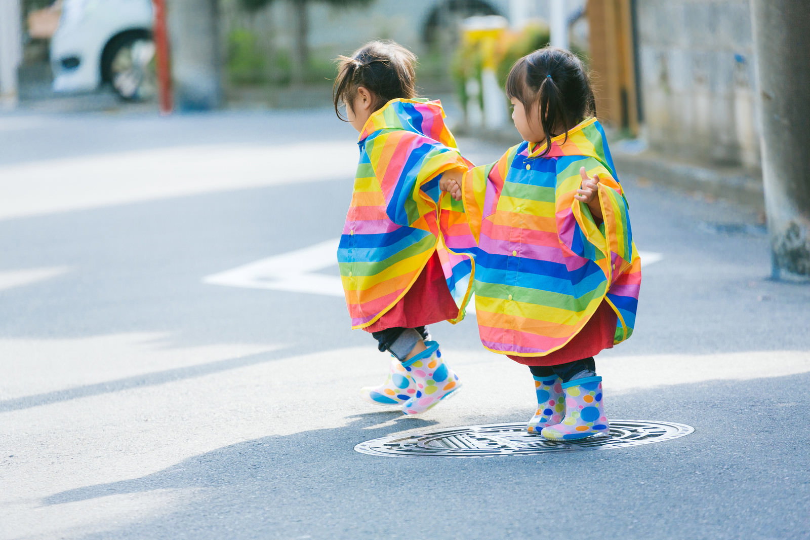 カラフルな雨合羽を着て手をつないで散歩する双子の女の子［モデル：あおみどり］