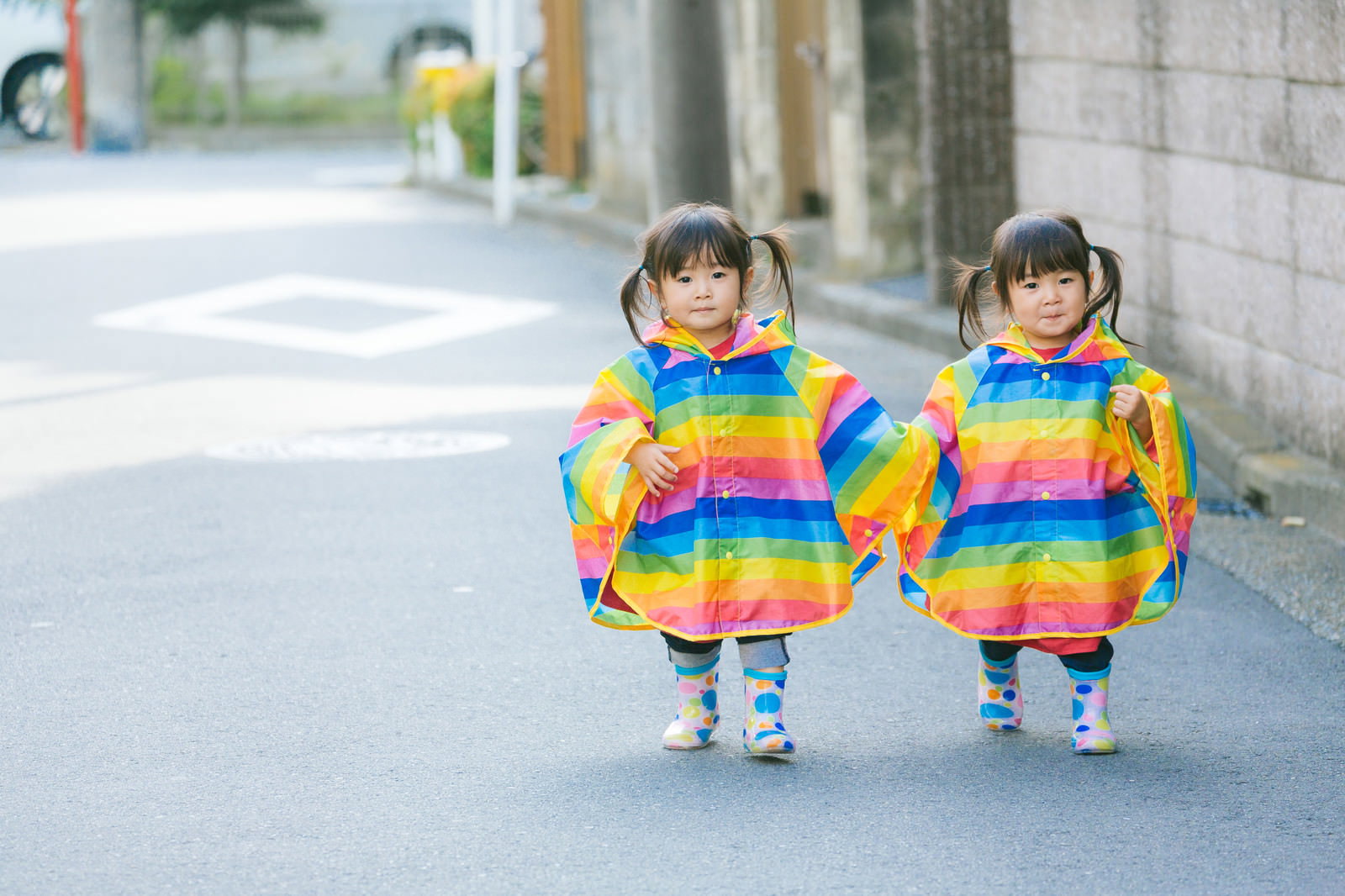 虹色のストライプ柄レインポンチョを着て手をつないで歩く双子の女児［モデル：あおみどり］