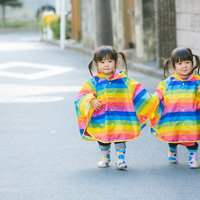仲良く手をつないで散歩するカラフルなレインポンチョ姿の双子の姉妹（2歳）の写真