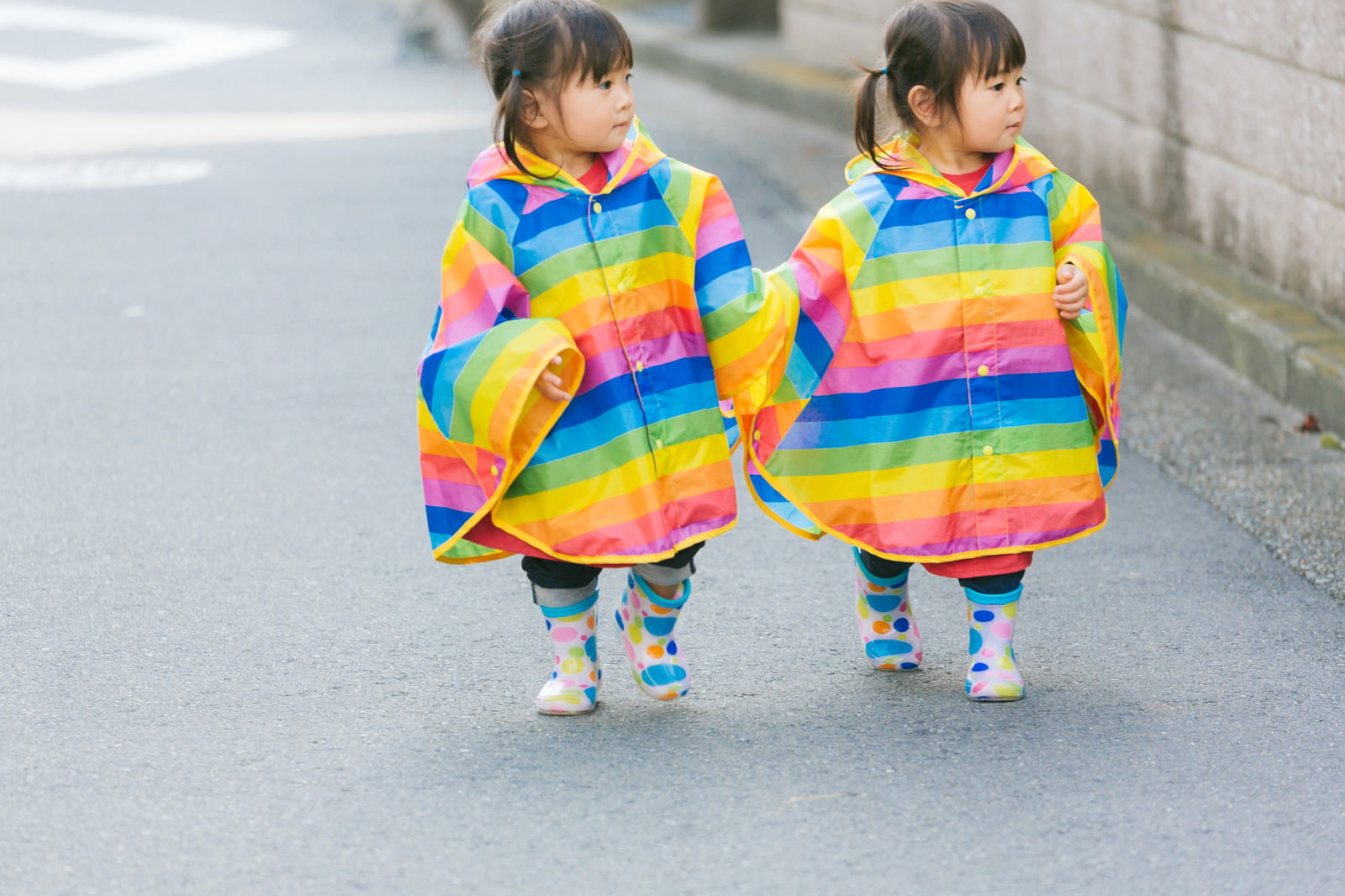 虹色のストライプ柄レインコートを着て手をつないで歩く双子の女の子［モデル：あおみどり］