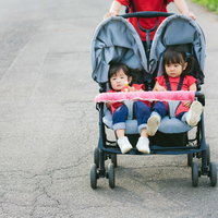 赤いヘルメットをかぶった双子の姉妹が乗った二人乗りベビーカーの写真