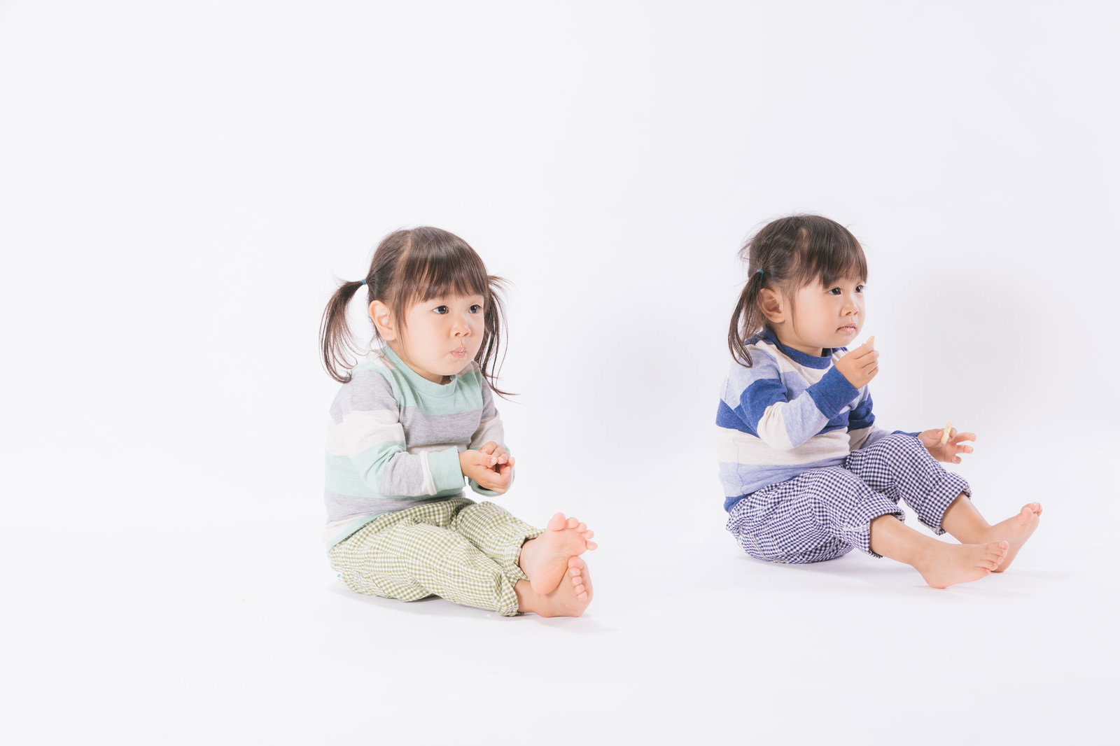 白い服と青い服を着た双子の女の子が床に座ってお菓子を食べている［モデル：あおみどり］