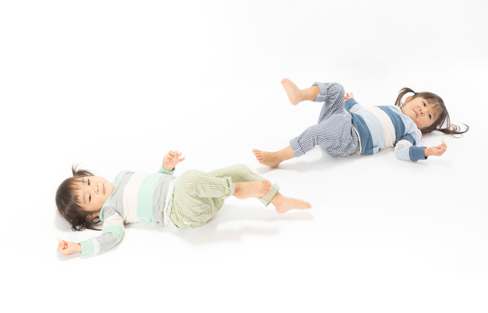 白い背景で仰向けに寝転がり足を上げてイヤイヤする双子の女の子［モデル：あおみどり］