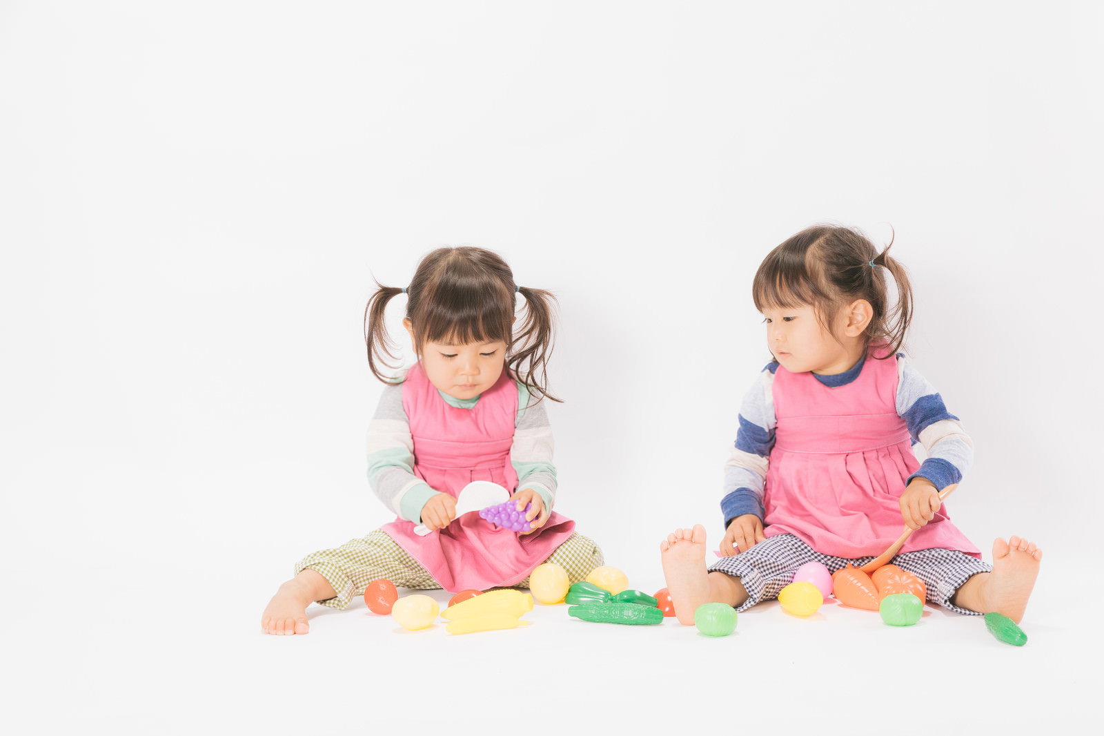 ピンクのエプロンを着けた双子の女の子がおままごとで遊んでいる様子［モデル：あおみどり］