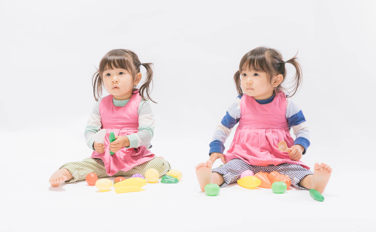 ピンクのエプロンを着た双子の女の子がおままごとで遊んでいる様子［モデル：あおみどり］