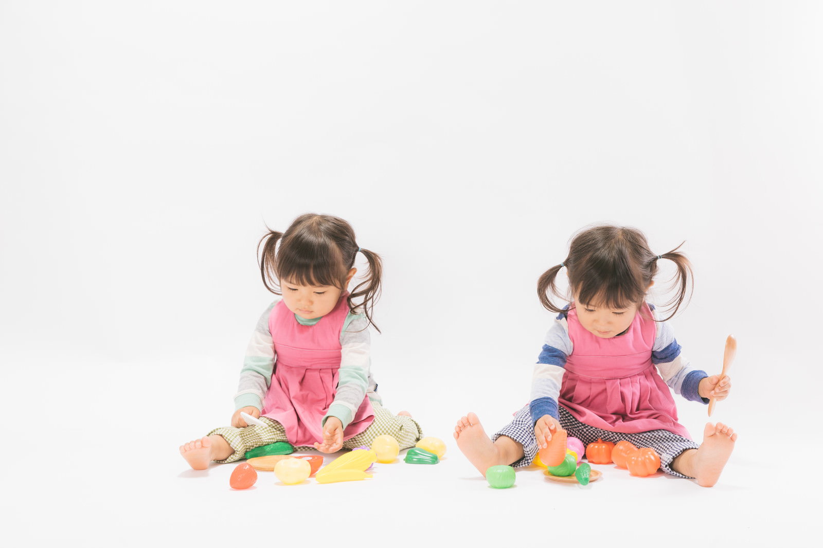 ピンクと紫のエプロンを着た双子の女児がツインテールで床におもちゃを広げておままごとをしている様子［モデル：あおみどり］