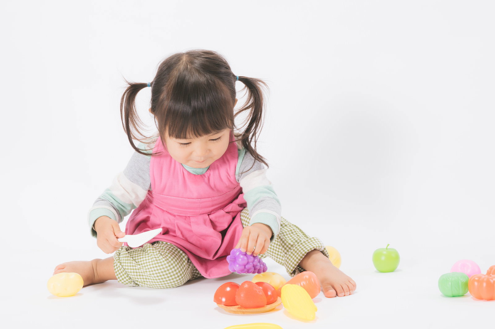 ピンクのエプロンを着けたツインテールの女の子がおもちゃのお皿におままごとの食べ物を盛り付けている［モデル：あおみどり］