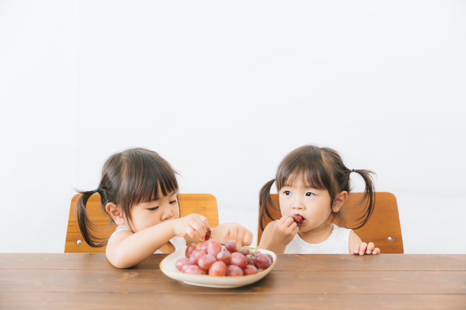 木製テーブルでお皿のぶどうを食べる双子の女の子［モデル：あおみどり］