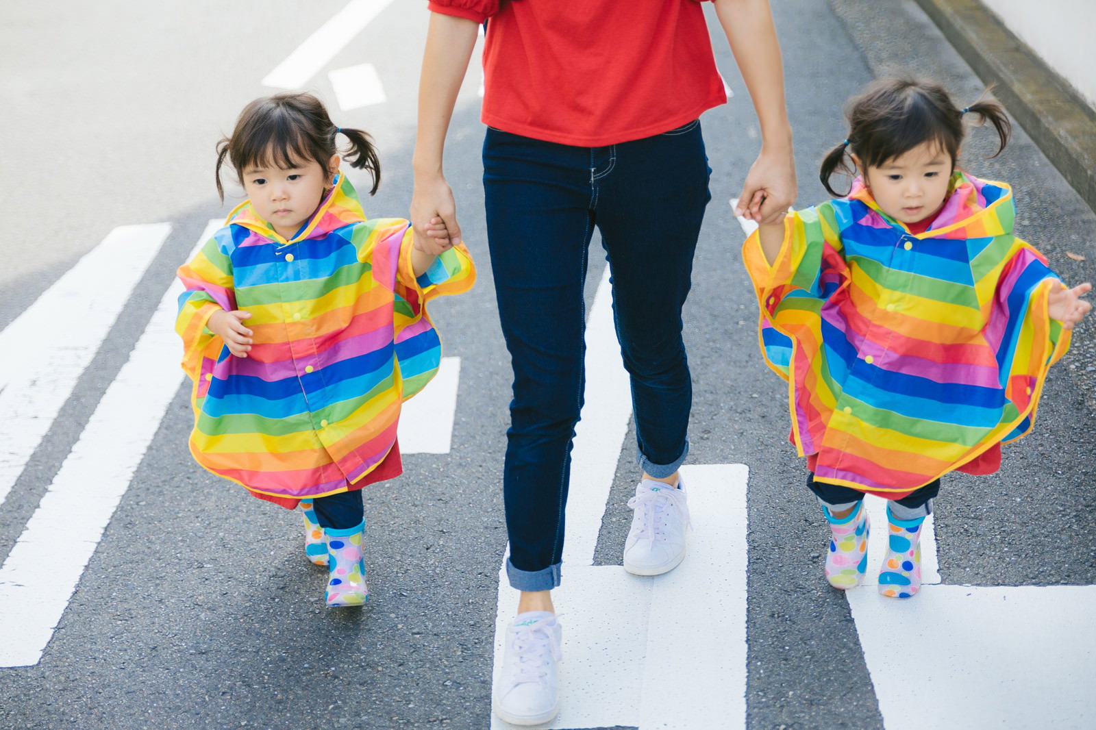 カラフルなボーダーシャツを着た双子の女の子が親と手をつないで公道を歩く様子［モデル：あおみどり］