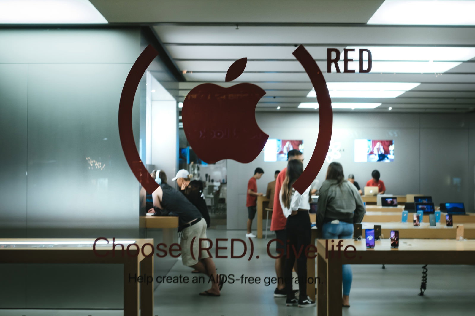 白い天井と壁に赤いAppleロゴが掲げられたストア内部。複数の来店客が木製の陳列台周辺に立っている